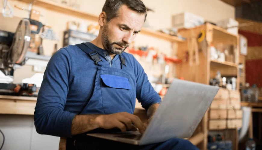 A man in blue overalls works on a laptop in a workshop with various tools and equipment on shelves in the background.