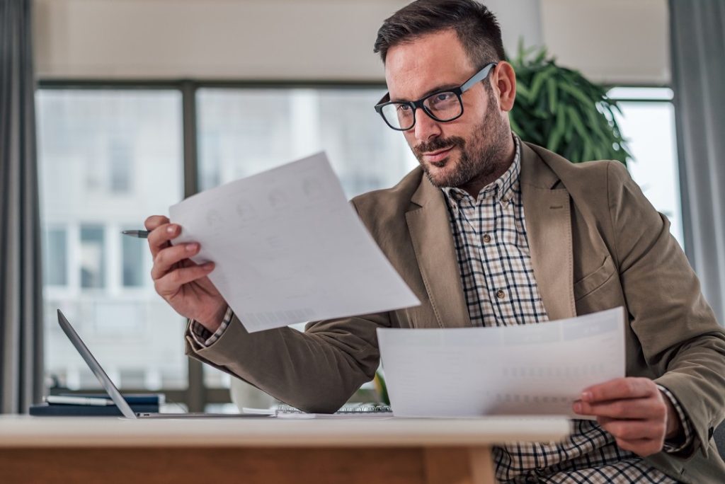 Man with glasses and a beard reviews documents at a desk, with a laptop and a plant in the background.