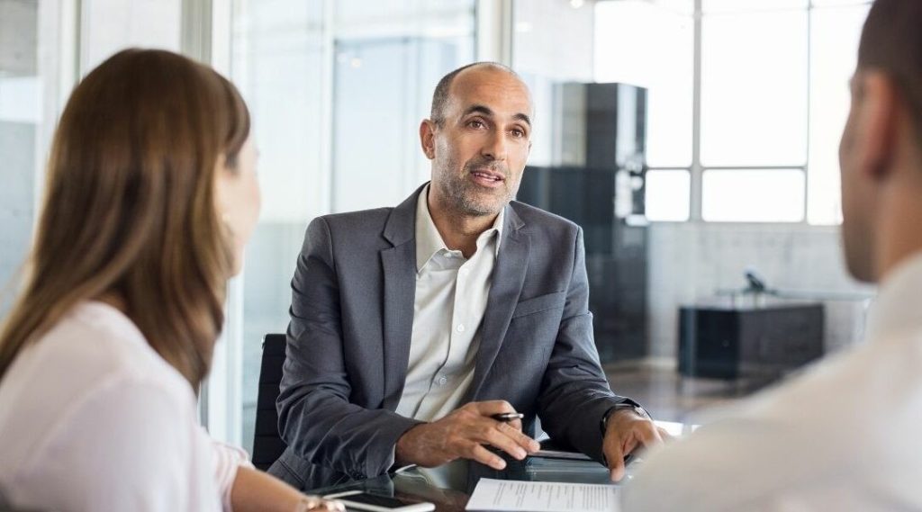 A businessman in a suit discusses paperwork with two colleagues in a modern office setting with glass walls.