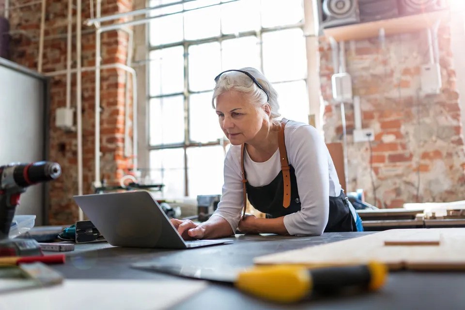 Older woman in an apron works at a laptop in a rustic workshop, surrounded by tools and equipment.