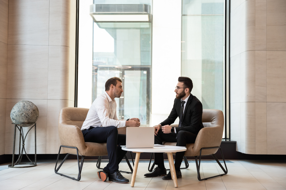 Two men in professional attire sit in armchairs and discuss business in a modern, bright office lobby with large windows.