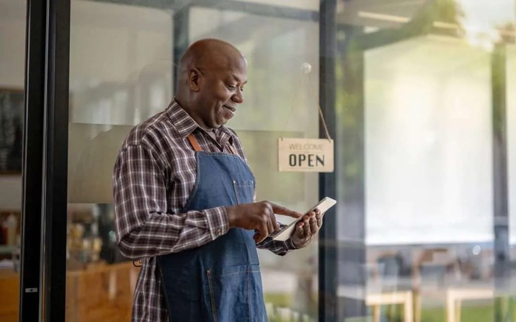 Man in plaid shirt and apron using a tablet inside a store with an "Open" sign on the glass door.