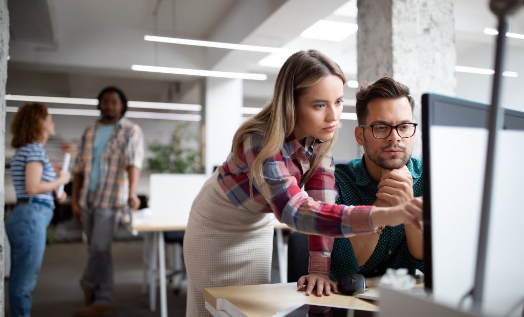 Two colleagues collaborate at a computer in a modern office, while two others converse in the background.