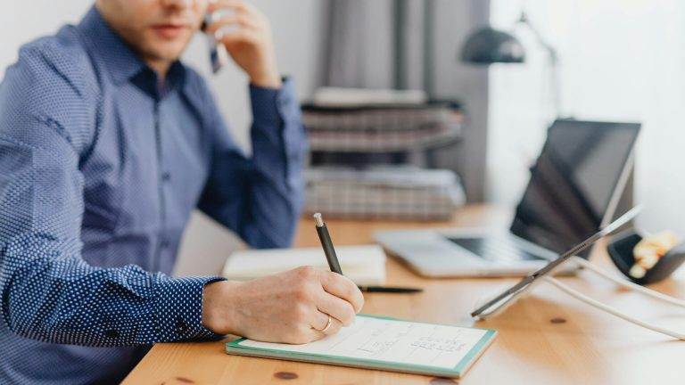 Person in a blue shirt writing in a notebook while on the phone, seated at a wooden desk with a laptop and tablet.
