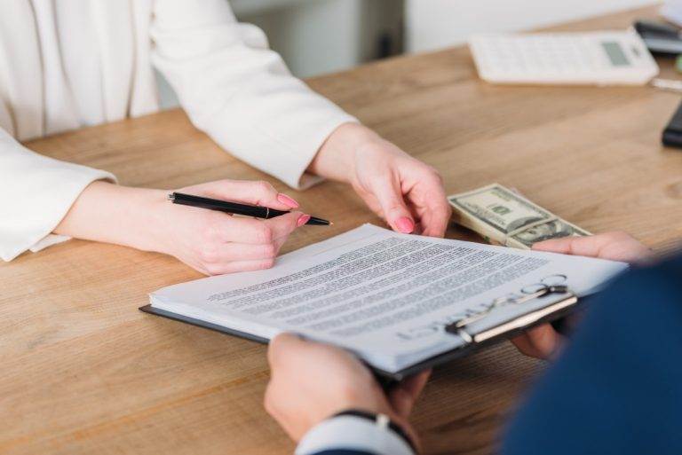 Two people exchange documents and cash on a wooden desk, with one person holding a pen and a calculator in the background.