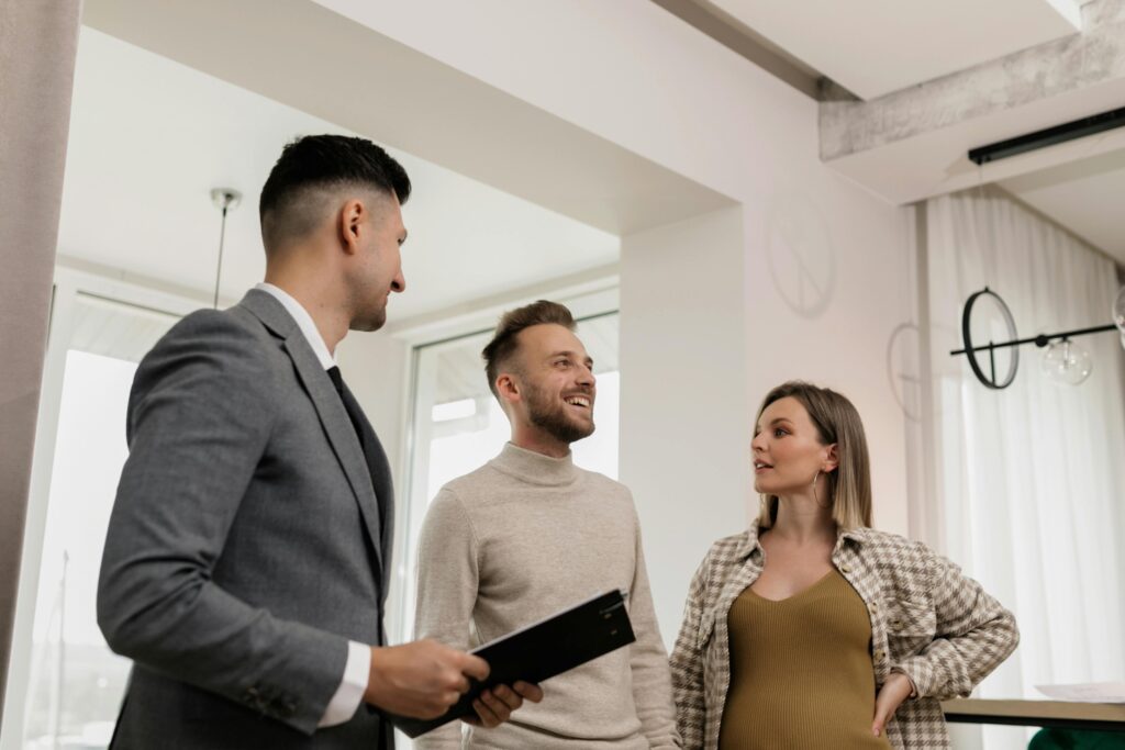 A man in a suit with a tablet talks to a smiling couple in a modern, well-lit room with large windows and stylish decor.