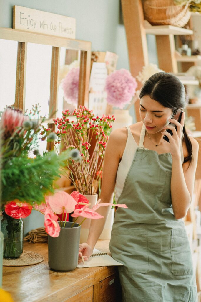 A florist in a gray apron talks on the phone, surrounded by vibrant flowers and taking notes in a cozy shop.