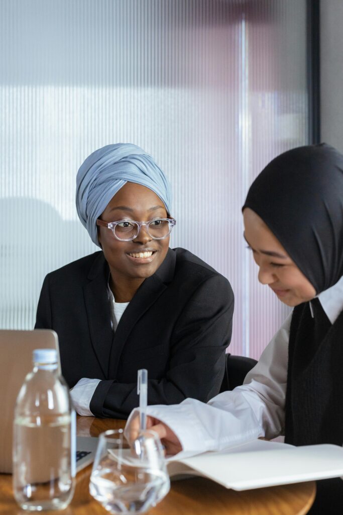 Two women in hijabs, one with a blue scarf and glasses, discuss at a table with a laptop, notebook, and water bottles.