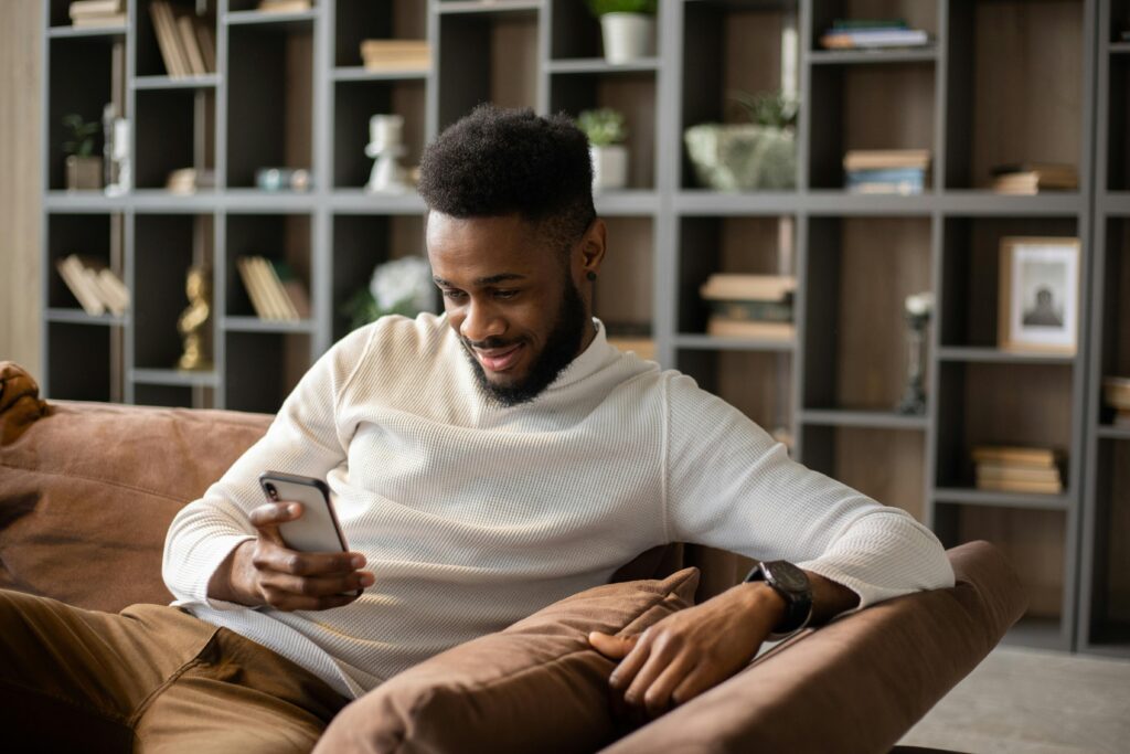 Man in a white sweater sitting on a brown couch, smiling while looking at his phone, with a bookshelf in the background.