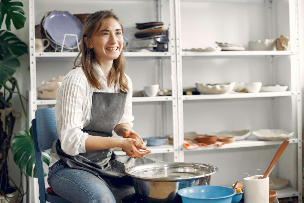 Smiling woman in an apron making pottery at a workbench, surrounded by shelves filled with ceramic pieces and plants.