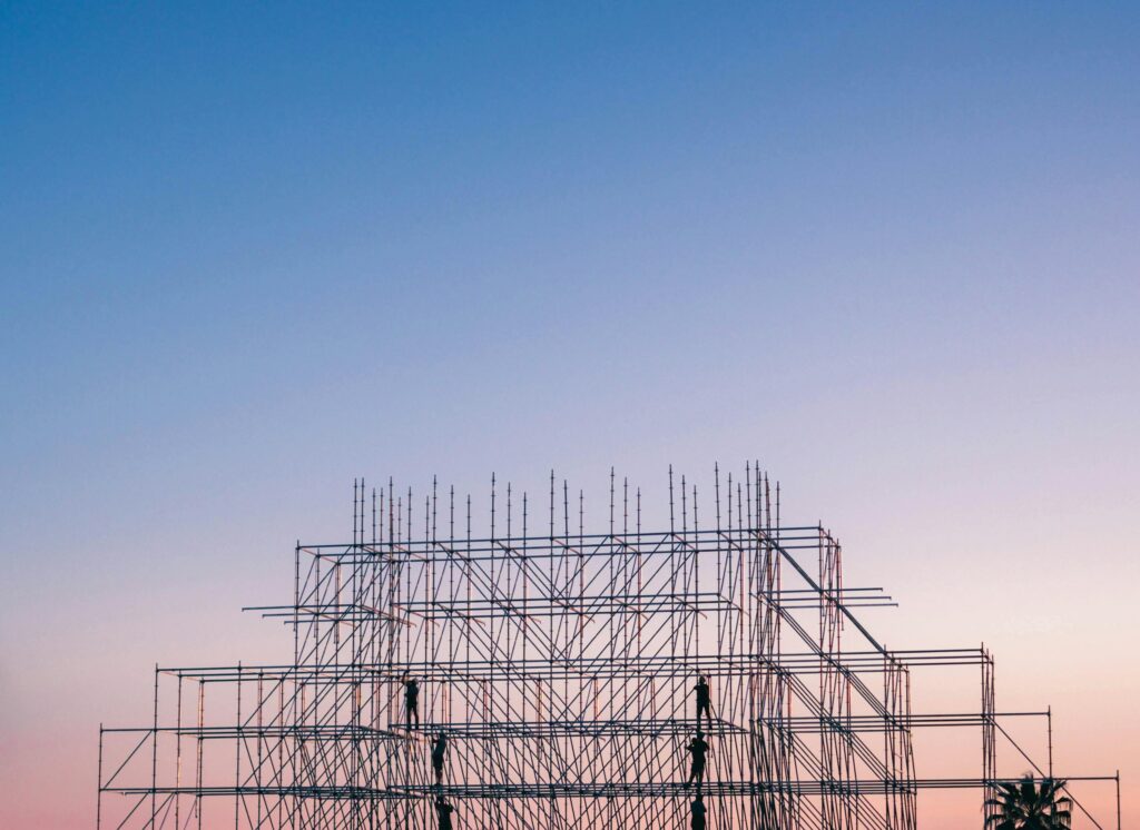 Workers climb a metal scaffolding structure under a clear blue and pink gradient sky at sunrise or sunset.