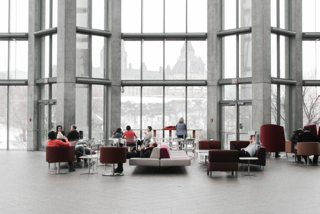 People sitting and conversing in a spacious, modern lobby with large floor-to-ceiling windows. An old building is seen outside.