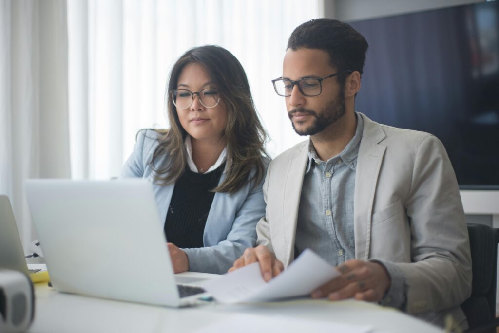 Two professionals in business attire work together at a laptop, reviewing documents in a bright office setting.