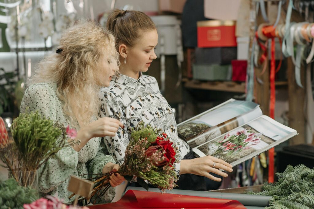 Two women standing in a flower shop, holding a floral arrangement and looking through a book of floral designs.