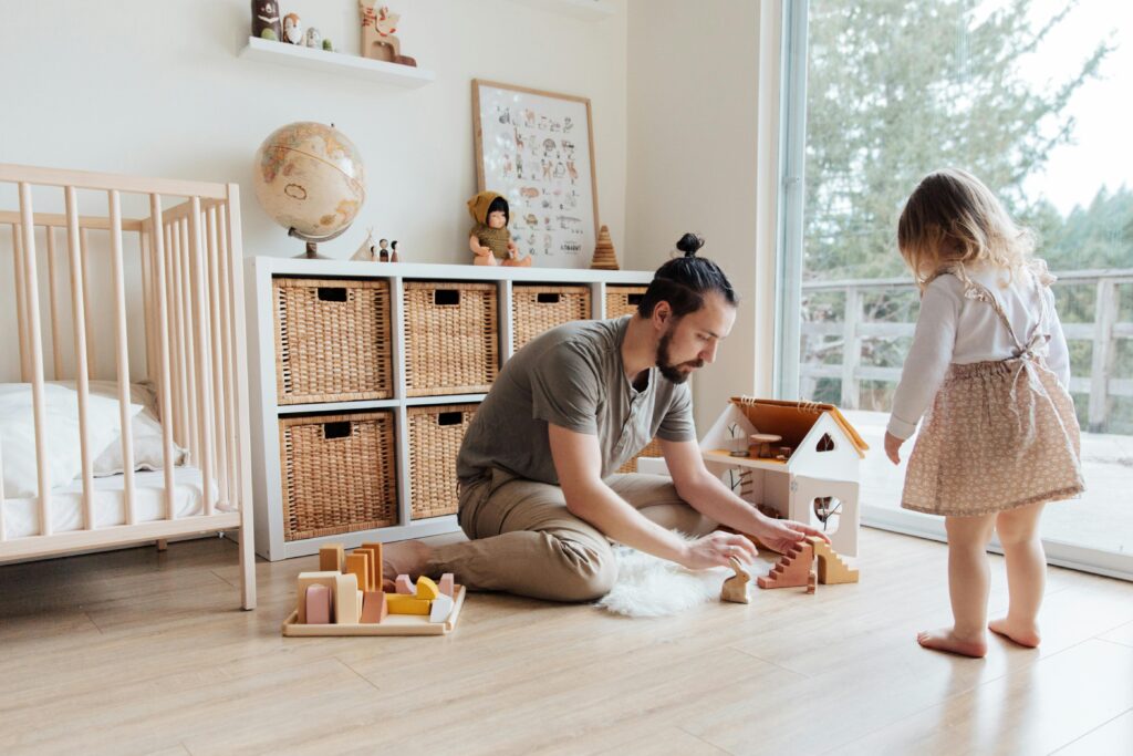 Man sitting on floor playing with wooden toys with a small child in a nursery, wicker baskets and a globe in the background.