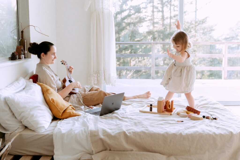 Woman with a ukulele sits on bed, watching a child play with wooden toys in a bright, airy room with a forest view window.