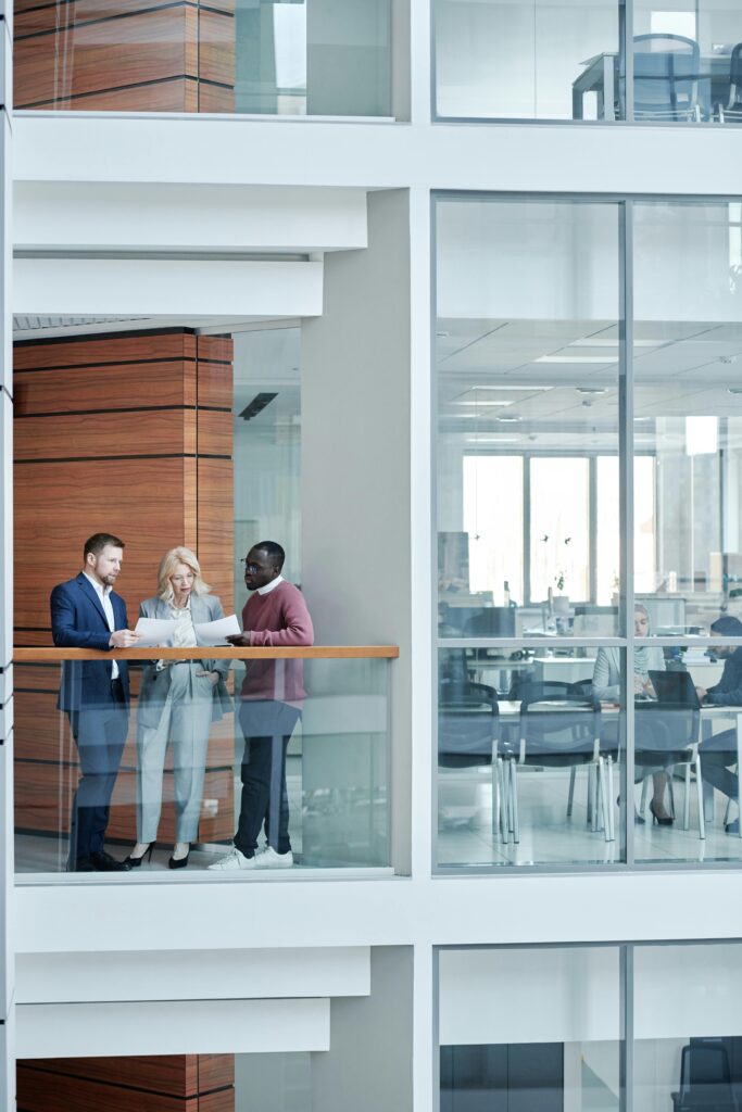 Three business professionals discuss papers on a modern glass-walled balcony, with an office setting visible in the background.