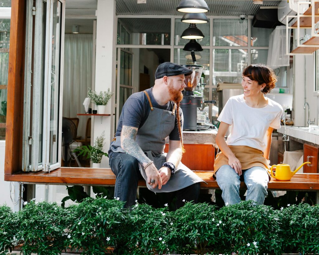 Two café workers sitting on a counter, chatting and smiling, with plants in the foreground and a coffee machine behind them.