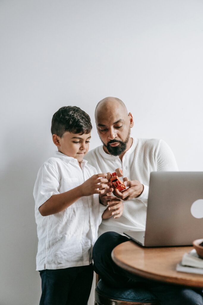 An adult and a child in white shirts examine a toy in front of a laptop at a wooden table against a white background.