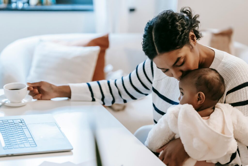 Woman holding a baby while sitting at a desk with an open laptop and holding a coffee cup, in a cozy home setting.