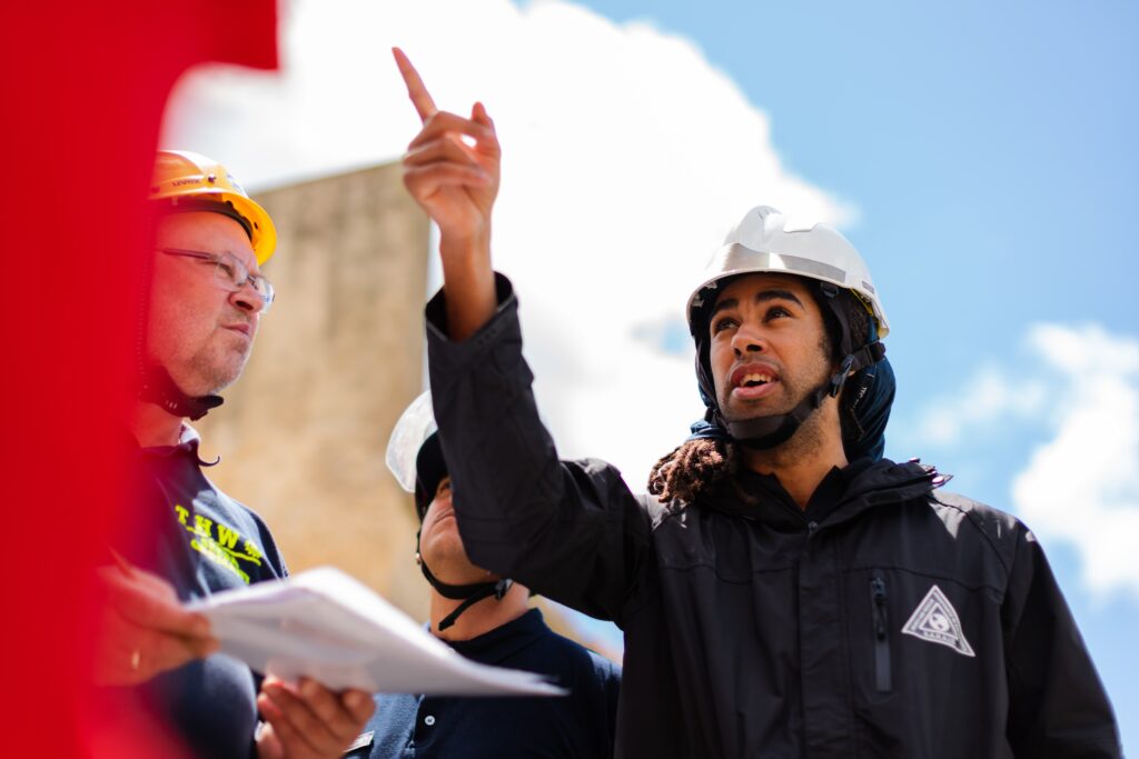 Construction workers in hard hats and reflective jackets discussing a project outdoors. One worker points upward.