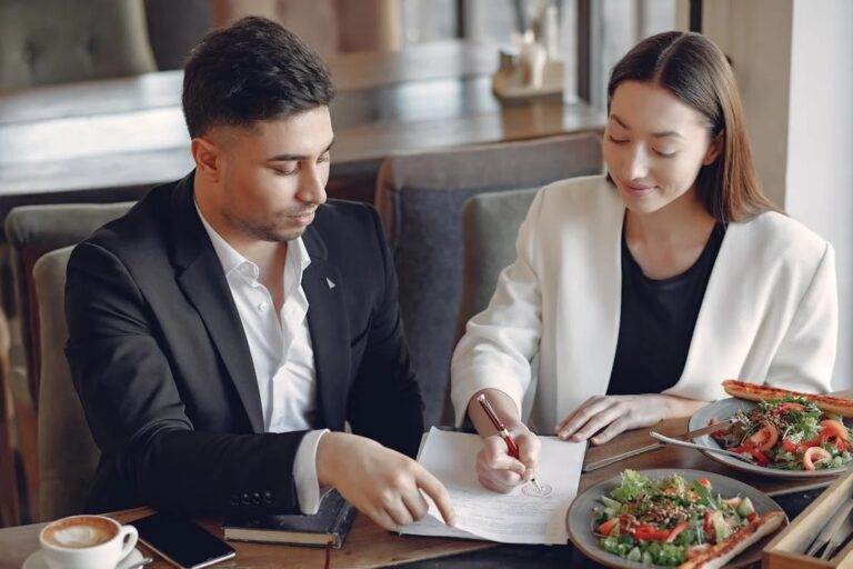Two professionals discussing documents over lunch, emphasizing the importance of avoiding fines for deceptive conduct when selling a business.