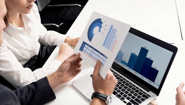 Two professionals discuss partnership valuation, analyzing charts and graphs on paper and a laptop with bar graphs.