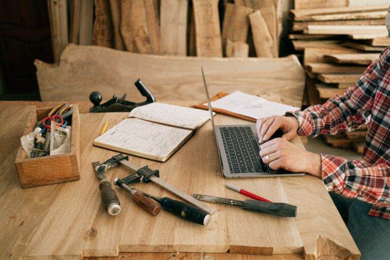 Person typing on laptop at a woodworking table, alongside tools and notes, discussing legal requirements for selling a manufacturing business.