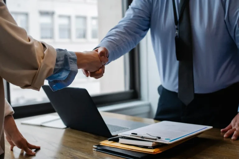 Two people shaking hands over a desk with a laptop and documents, symbolizing a business agreement in NSW.