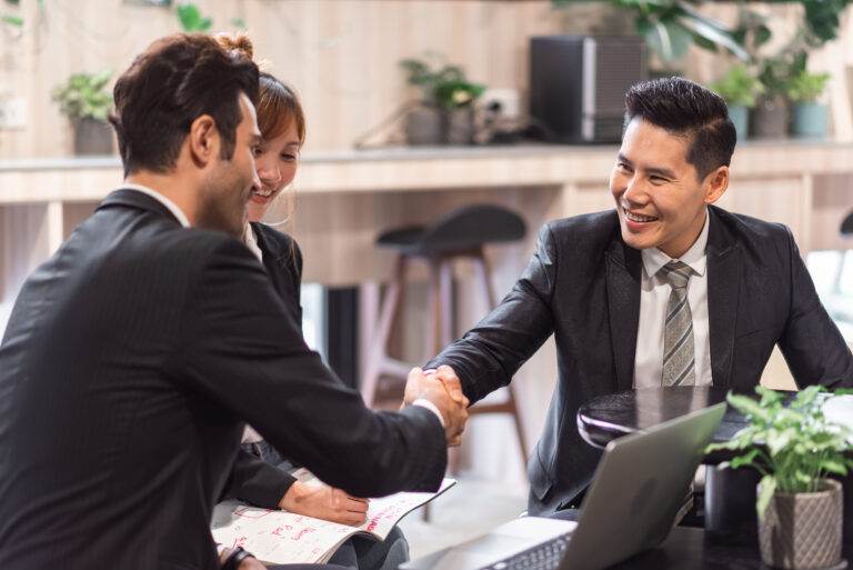 Three professionals in suits discuss how to calculate goodwill, shaking hands in an office with a laptop and plants.
