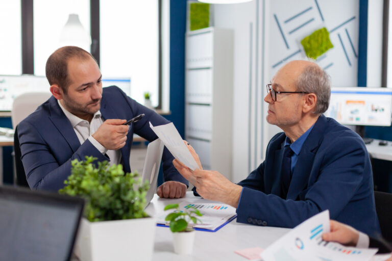 Two businessmen in suits discuss documents related to an earn-out clause in a modern office setting.