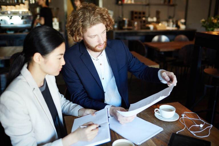Two business professionals discuss escrow holdback terms during a business sale meeting in a cafe.